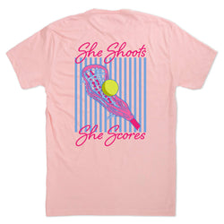 A light pink short-sleeve t-shirt shown flat against a white background, featuring a large graphic print on the back with the text 'She Shoots She Scores' in pink script, a pink lacrosse stick, and a yellow lacrosse ball over a blue and pink striped background.