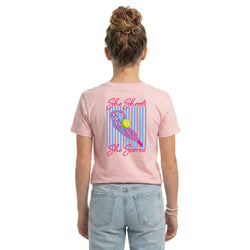 A woman shown from the back wearing a light pink t-shirt with a lacrosse-themed graphic print that reads 'She Shoots She Scores' alongside a lax stick and ball, paired with light blue jeans against a plain white background.