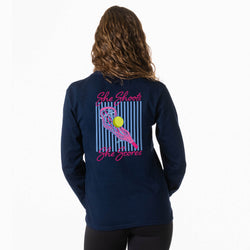A woman viewed from behind wearing a navy long-sleeve shirt with a colorful back graphic featuring a pink lacrosse stick, a yellow lacrosse ball, and pink text reading 'She Shoots The Scores' against a white background.
