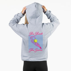 A person shown from the back wearing a light heather gray hoodie with a colorful graphic on the back. The graphic features pink text reading 'She Shoots She Scores' around a pink lacrosse stick and yellow lacrosse ball over blue vertical stripes.