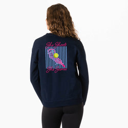 A woman viewed from the back wearing a navy blue sweatshirt with a colorful tennis-themed graphic on the back. The graphic features pink text reading 'She Shoots The Scores' and a pink lacrosse stick holding a yellow ball over blue vertical stripes.