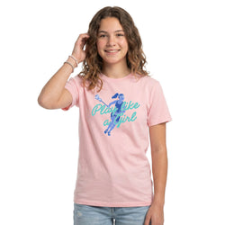 A young woman with wavy brown hair smiling and posing in a light pink t-shirt with a blue graphic of a lacrosse player and the text 'Play like a girl'. She is wearing light blue jeans against a solid white background.