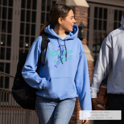 A woman walking outdoors wearing a light blue hoodie with a teal graphic and the words 'Play like a girl' across the front. She is also wearing blue jeans and carrying a black backpack.