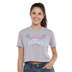 A smiling young woman with her hair in a ponytail models a light heather gray cropped t-shirt featuring a lacrosse graphic that reads 'pretty TOUGH' in pink and blue. She is wearing blue jeans against a solid white background.