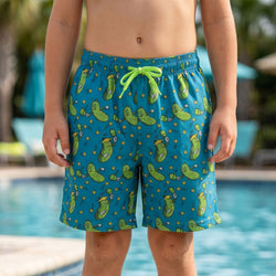 Child wearing green swim shorts with green pickle pattern by a pool.