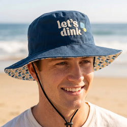 Person wearing a blue bucket hat with 'let's dink' text on a beach