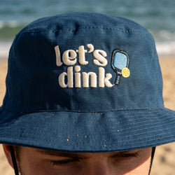 Navy blue bucket hat with 'let's dink' text and paddle ball graphic on a beach.