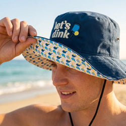 Person wearing a navy bucket hat with 'let's dink' text on a beach