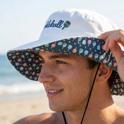 Person wearing a white bucket hat with colorful paddle pattern on a beach