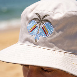 Beige bucket hat with pickleball paddles and palm tree design on a beach.