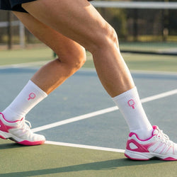 Person wearing white socks with pink pickleball accents and tennis shoes on a tennis court.
