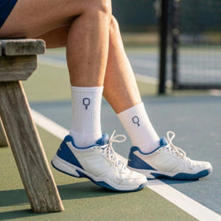 Person sitting on a tennis court wearing white and blue sneakers with socks featuring a pickleball paddle logo.