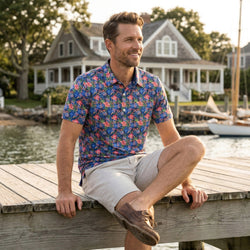 Man sitting on a dock by a lake wearing a colorful floral and pickleball patterned polo shirt and beige shorts.