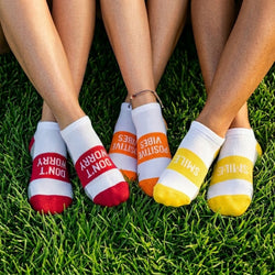 Three pairs of colorful socks with text on the toes, worn by people sitting on grass.