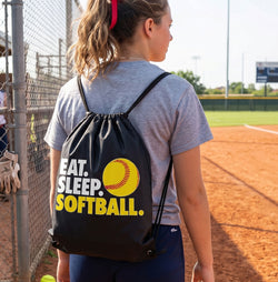 Person wearing a drawstring bag with 'Eat. Sleep. Softball.' text on a baseball field