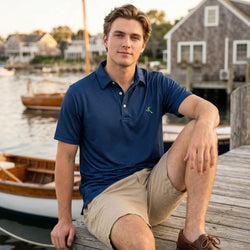Man in a blue soccer polo shirt and beige shorts sitting on a dock with boats and houses in the background.