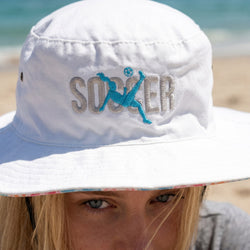 White bucket hat with 'SOCCER' and a soccer player graphic on a beach.