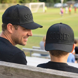 Man and child wearing black 'SOCCER' hats at a sports event.