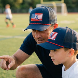 Man and young boy wearing matching soccer caps with American flag design on a sports field.