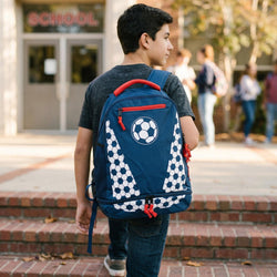 Child with a blue backpack featuring a soccer ball design, standing outside a school.
