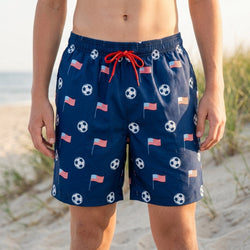 Person wearing navy blue swim shorts with American flag and soccer ball pattern on a beach.