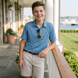 Young man in a blue soccer patterned polo shirt and beige shorts standing on a porch with a scenic background.
