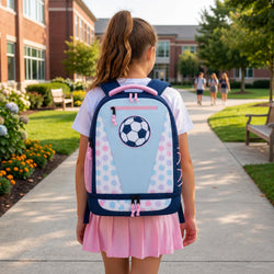 A young girl viewed from behind wearing a light blue and pink soccer-themed backpack on a sunny school walkway.