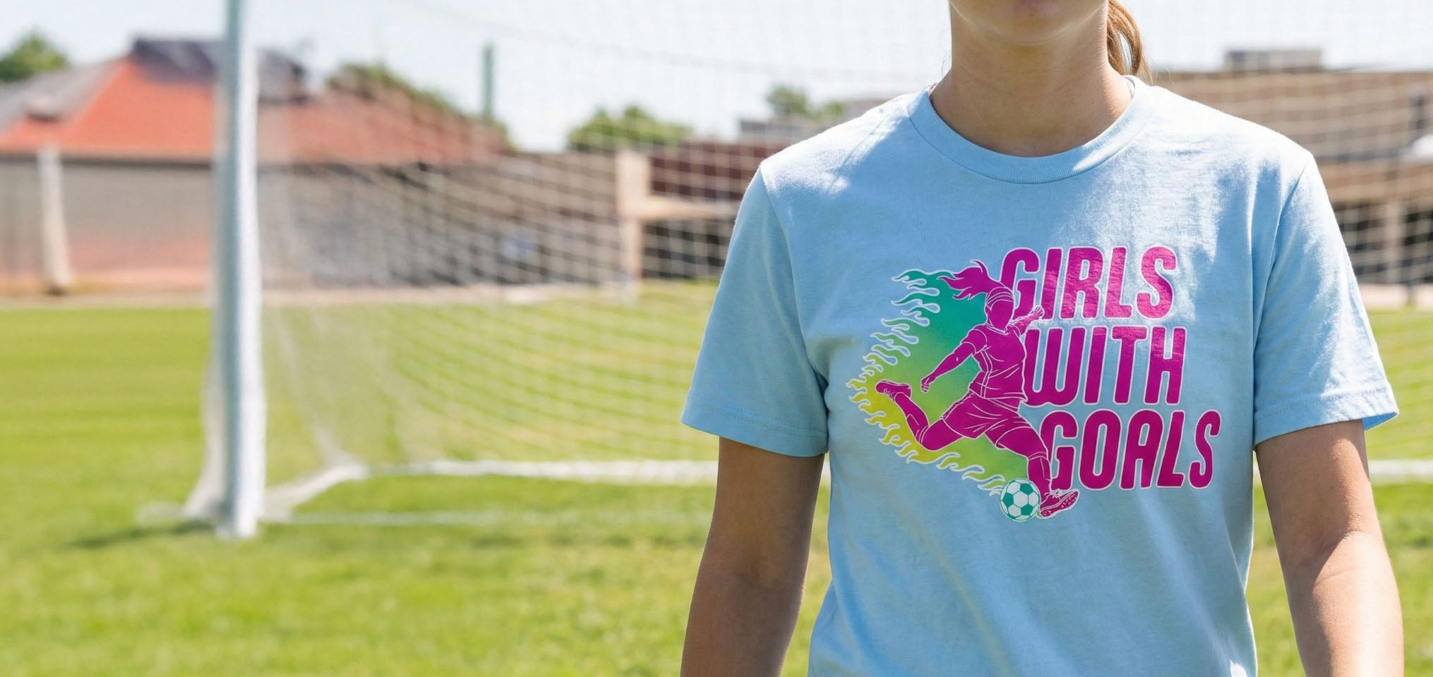Girl wearing light blue soccer t-shirt featuring pink Girls With Goals slogan and player silhouette.