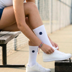 Person wearing white socks with a blue tennis logo and white sneakers, sitting on a wooden bench.