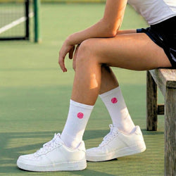 Person sitting on a tennis court wearing white sneakers and socks with pink tennis ball.