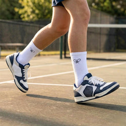 Person wearing white socks with a tennis logo and navy blue and white tennis shoes on a tennis court.