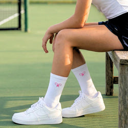 Person sitting on a bench wearing white sneakers and socks with a pink tennis design on a tennis court.