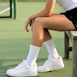 Person sitting on a bench wearing white sneakers and socks with a yellow tennis logo on a tennis court.