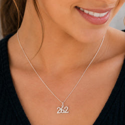 A close-up of a woman wearing a delicate silver chain necklace with a '26.2' cursive pendant, styled with a black knit top.