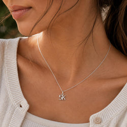 A close-up of a woman wearing a delicate silver chain necklace with a small '5K' initial pendant, styled with a cream-colored knit cardigan.
