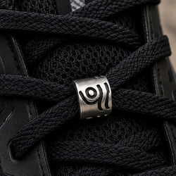 A close-up of a black athletic shoe with thick black laces threaded through a silver metal lace lock featuring a carved circular symbol.