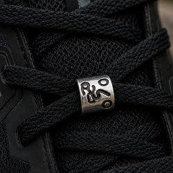 A close-up of black athletic laces threaded through a black shoe, with a silver metal eyelet featuring an engraved cycling figure.