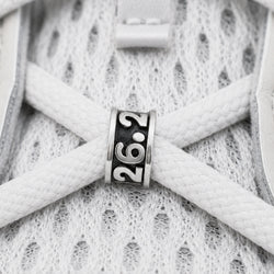 A close-up of a silver metal shoe charm with a black center and the numbers '26.2' engraved on it, resting on white athletic shoe laces over a white mesh sneaker upper.