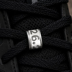 A close-up of a black sneaker with thick black laces threaded through a polished silver metal eyelet engraved with the numbers '26.2'.