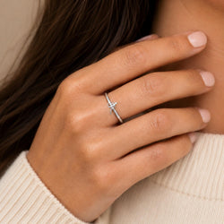 A close-up of a delicate silver ring with a small runner design, worn on a woman's finger. The ring is shown on a hand with light pink manicured nails, wearing a cream-colored ribbed sweater against a soft beige background.