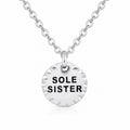 A silver disc pendant necklace with the words 'SOLE SISTER' engraved in black letters, suspended from a silver cable chain on a white background.