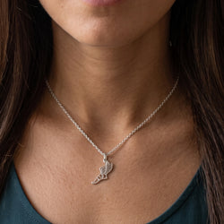 A close-up of a woman wearing a delicate silver chain necklace with a small silver pendant shaped like a stylized leaf or flame, shown against her neck and upper chest.