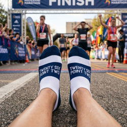 Person wearing 'Twenty Six Point Two' socks at a race finish line with runners in the background.