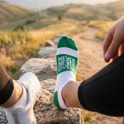Person wearing green socks with 'Never Never Give Up' text, sitting on a rock with a scenic background.