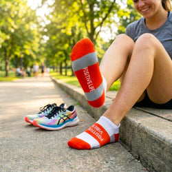 Person sitting on a park bench wearing orange and white socks with 'POSITIVE VIBES' & 'POSITIVE LIFE' text, next to colorful running shoes.