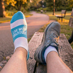 Person wearing blue and white socks with 'Live with Gratitude' text, sitting on a wooden bench in a park.