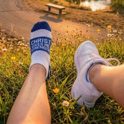 Person wearing socks with 'Christ Gives Me Strength' text, sitting on grass with a scenic background.
