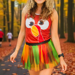 A woman wearing a turkey-themed costume top and a colorful tulle tutu skirt, standing on a leaf-covered path in a forest.