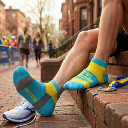 Person wearing blue and yellow running socks with Boston 26.2 text, sitting on a brick ledge with a running event in the background.