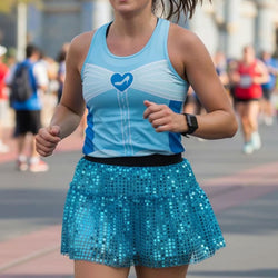 A woman running outdoors wearing a light blue athletic tank top with a blue heart and shoe graphic on the chest, paired with a bright blue sequined tutu skirt.